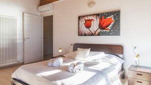 A neatly made bed with rolled towels, a wooden headboard, a nightstand with a lamp, and a painting of red flowers on the wall. - Apartment for the whole family. (Vercana)