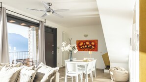 A modern living room with a sofa, dining table, and chairs. There is a ceiling fan, a painting on the wall, and a view of the outdoors. - Apartment for the whole family. (Vercana)