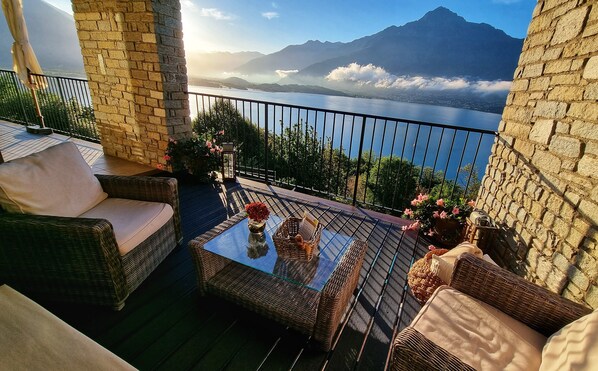 A balcony with wicker furniture, a glass table, and a view of a lake and mountains.