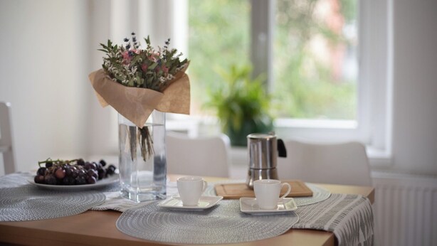 A table with a vase of flowers, a plate of grapes, and two white cups on a placemat.