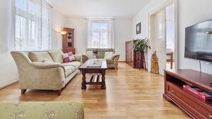 A spacious living room with a sofa, armchair, and coffee table. There is a TV on the right and a cabinet with a plant.