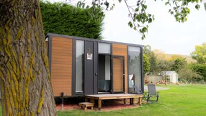 A modern, compact cabin with a wooden exterior and large glass windows, situated in a grassy backyard with a bench and chairs outside. - Mona Vale Country Estate by Tiny Away (Waikari)