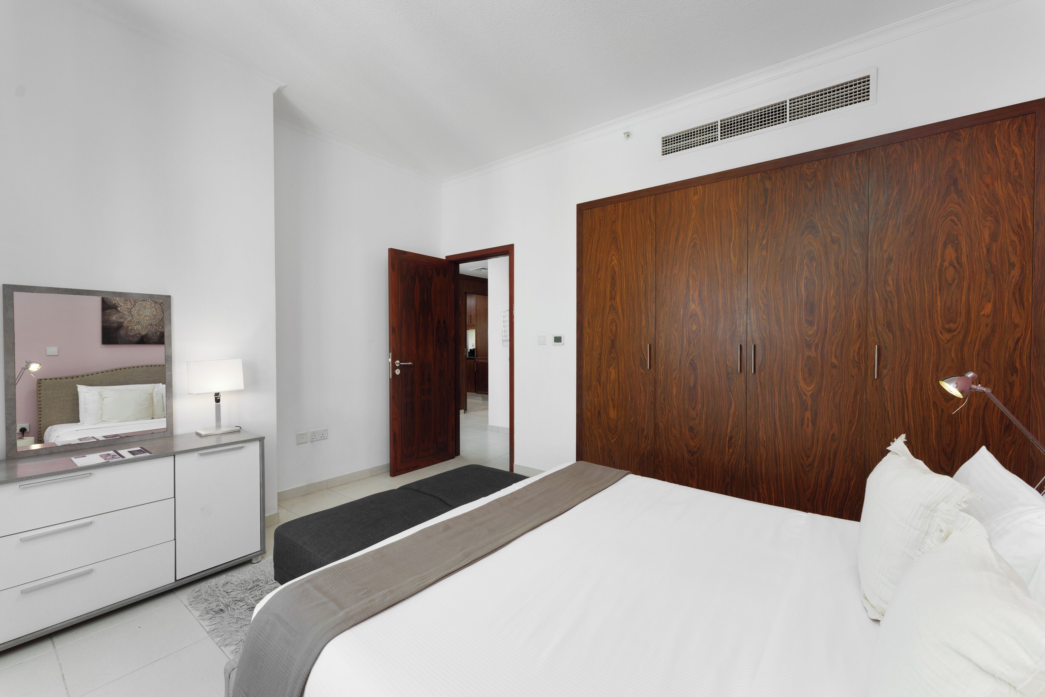 Un dormitorio moderno con un amplio armario de madera, una mesita de noche con lámpara y una cama con ropa de cama blanca.