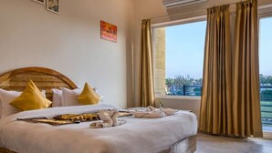 Quarto de hotel com uma cama grande, cabeceira de madeira e vista da cidade através de uma janela com cortinas.