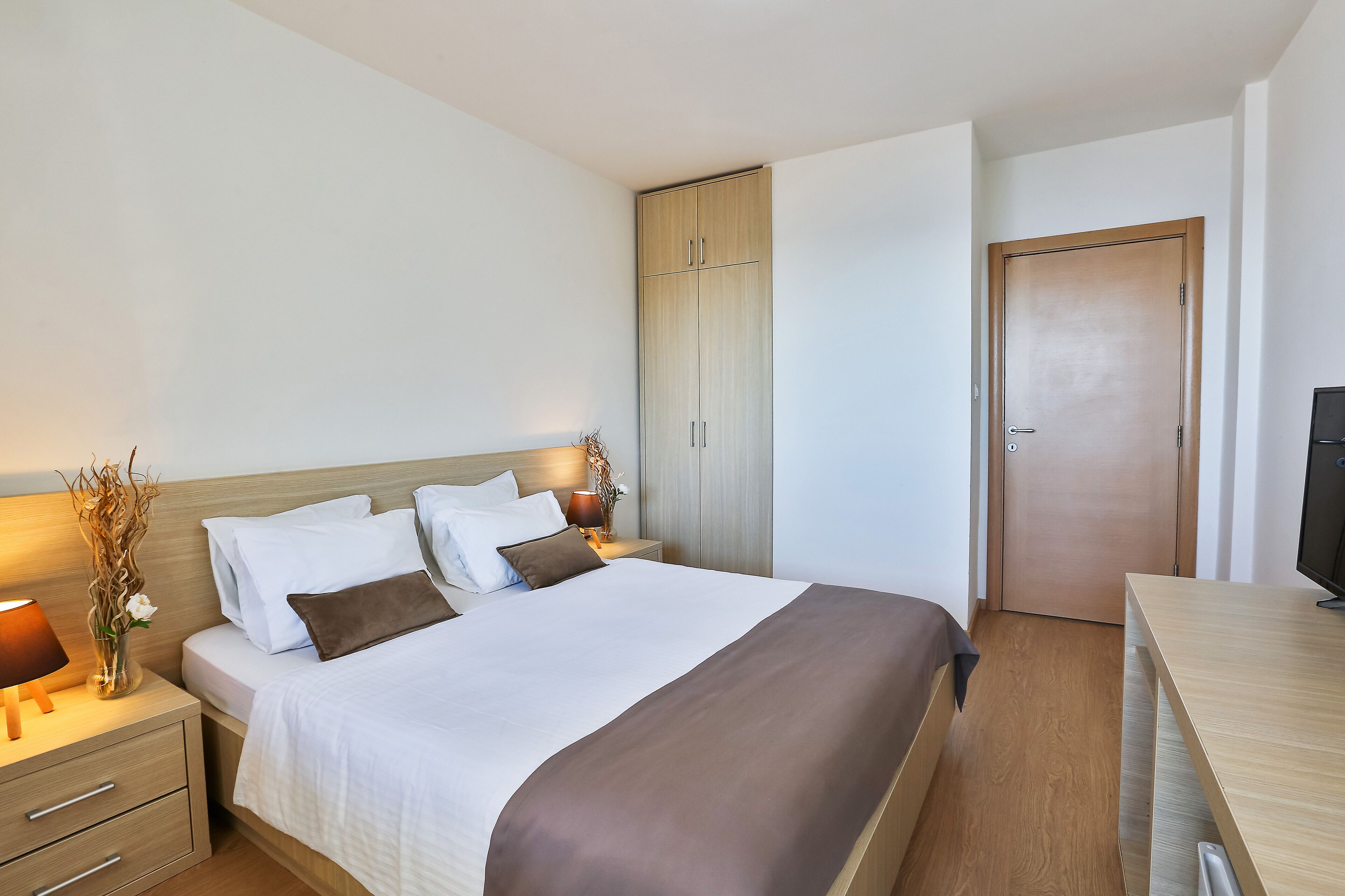 Una cama bien hecha con ropa de cama blanca y marrón, una mesita de noche con una lámpara y un armario de madera.