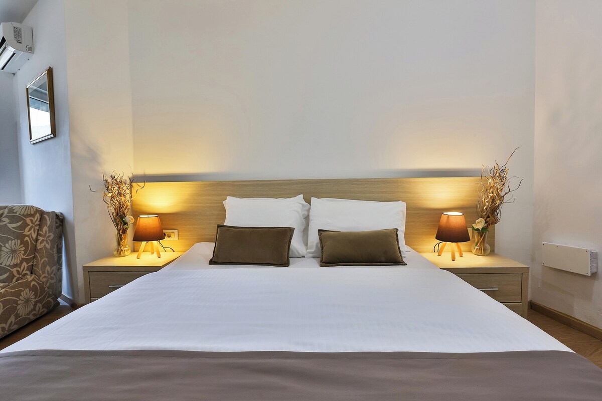 Una cama bien hecha con ropa de cama blanca y marrón, flanqueada por dos mesitas de noche con lámparas y pequeños jarrones.