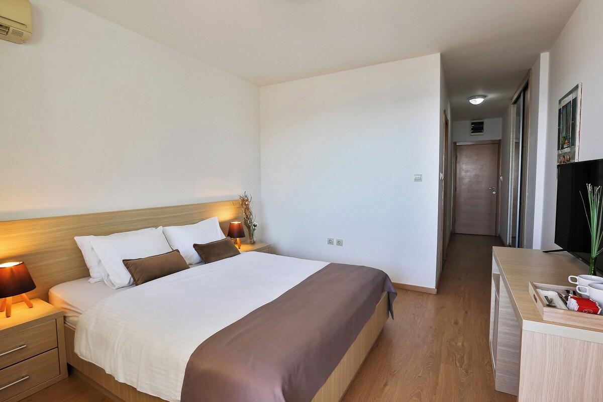 Una habitación de hotel con una cama bien hecha, lámparas de noche, una mesita de madera y un escritorio con una planta y una taza.