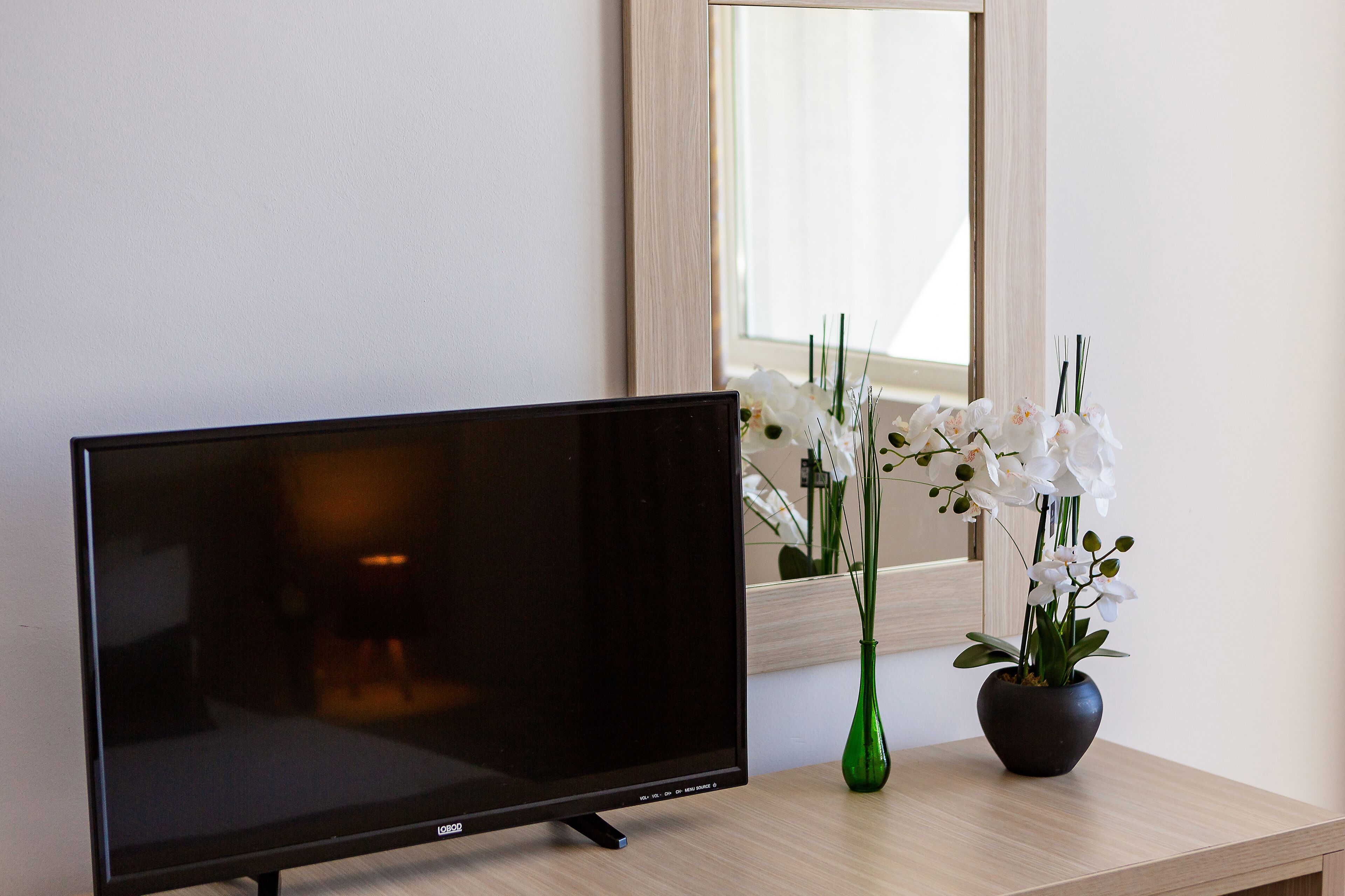 A flat-screen TV on a wooden table with a green vase of white flowers.