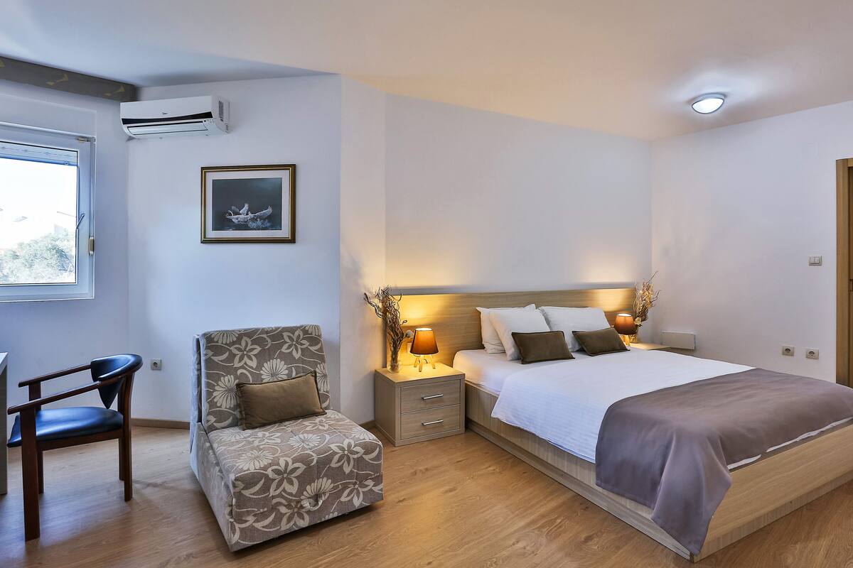 Un dormitorio con una cama, mesitas de noche, una silla, aire acondicionado, un cuadro enmarcado y una ventana.