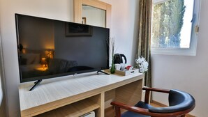 Habitación moderna con una televisión de pantalla plana grande sobre un escritorio de madera, una silla, una mesita con un florero y una ventana con vistas a árboles.