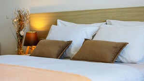 Una cama bien hecha con almohadas blancas y marrones, un cabecero de madera y una lámpara con un jarrón de flores secas a su lado.