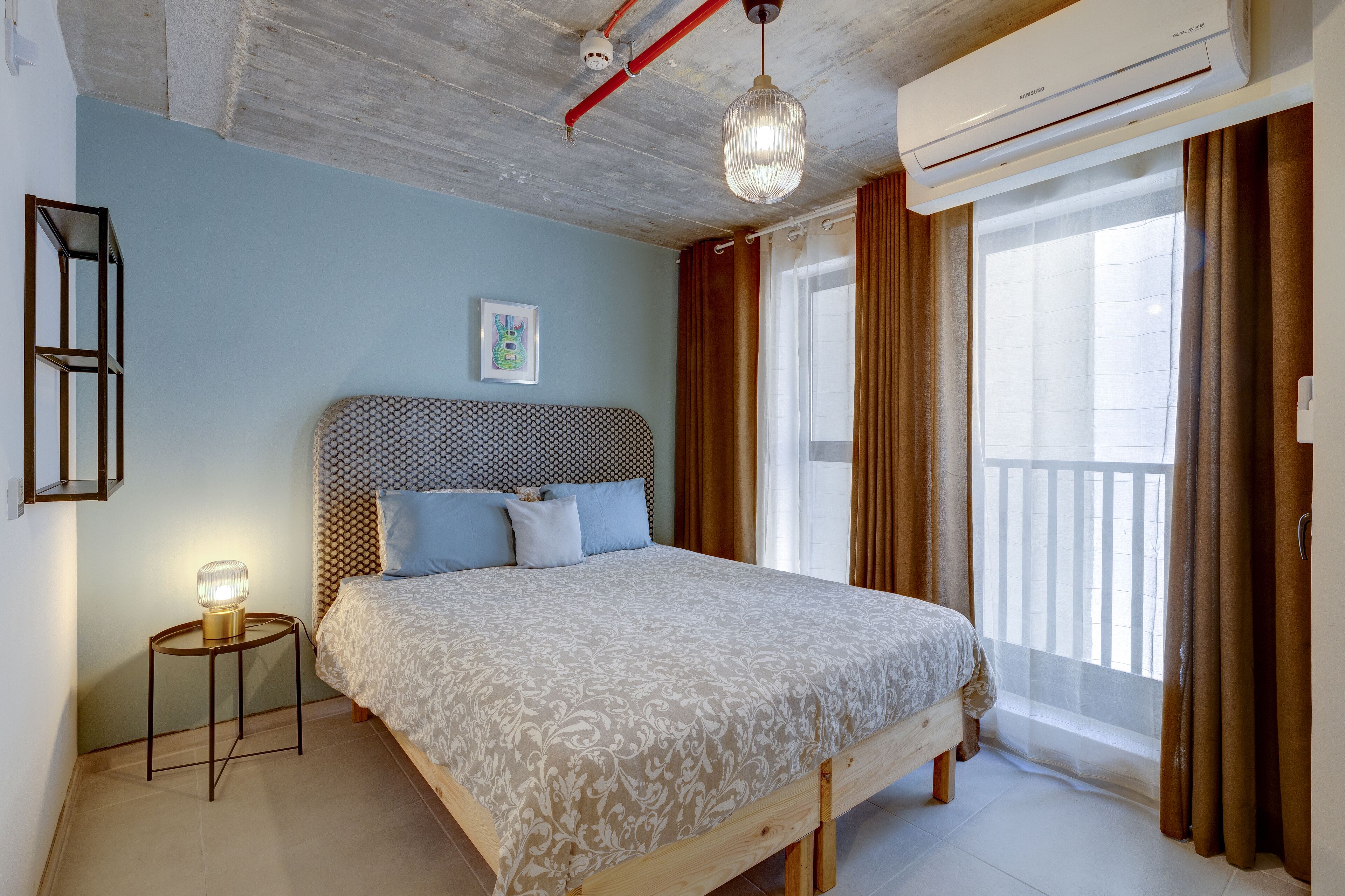 A bedroom with a bed, a bedside lamp, a window with curtains, and an air conditioning unit.