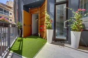 Un estrecho pasillo exterior con césped artificial verde, paneles de madera y macetas grandes con flores.