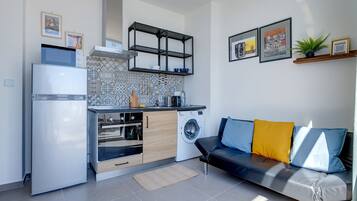 Una cocina compacta con refrigerador, horno y lavadora, un sofá con cojines, y una pared decorada con cuadros.