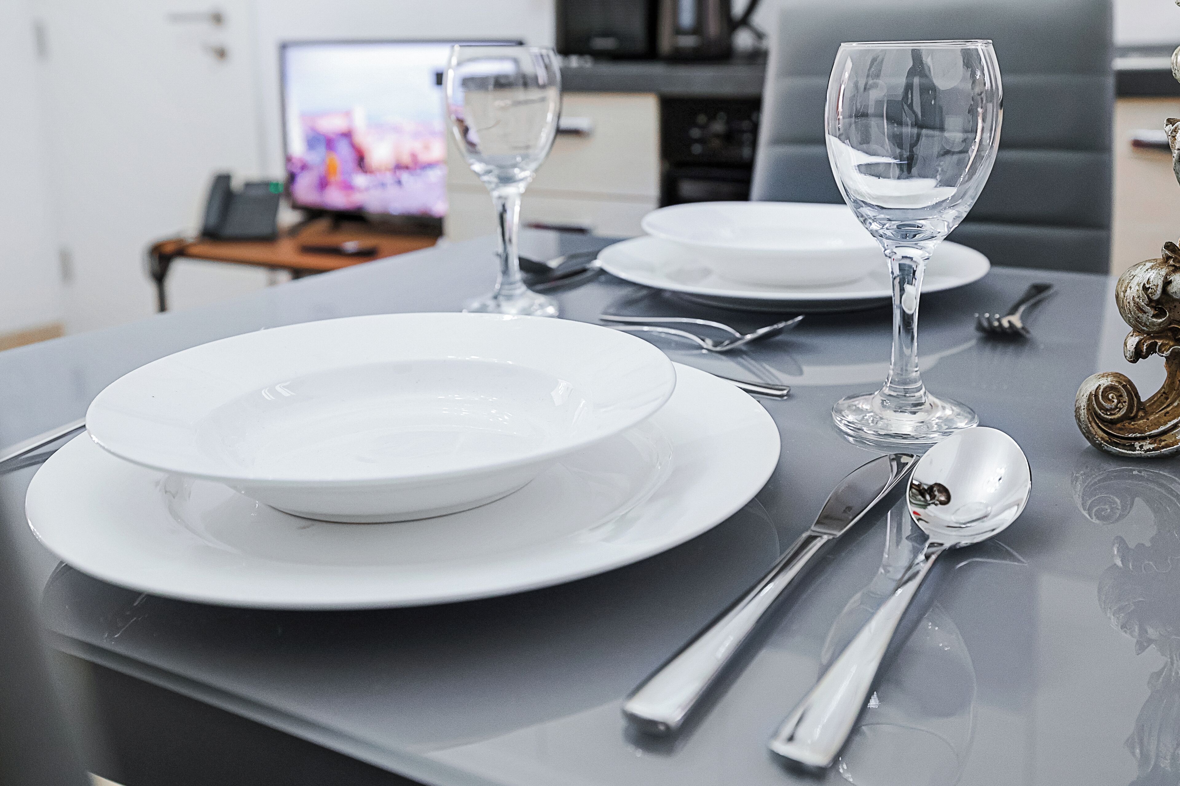 A dining table set with white plates, silverware, and clear wine glasses, with a television in the background.