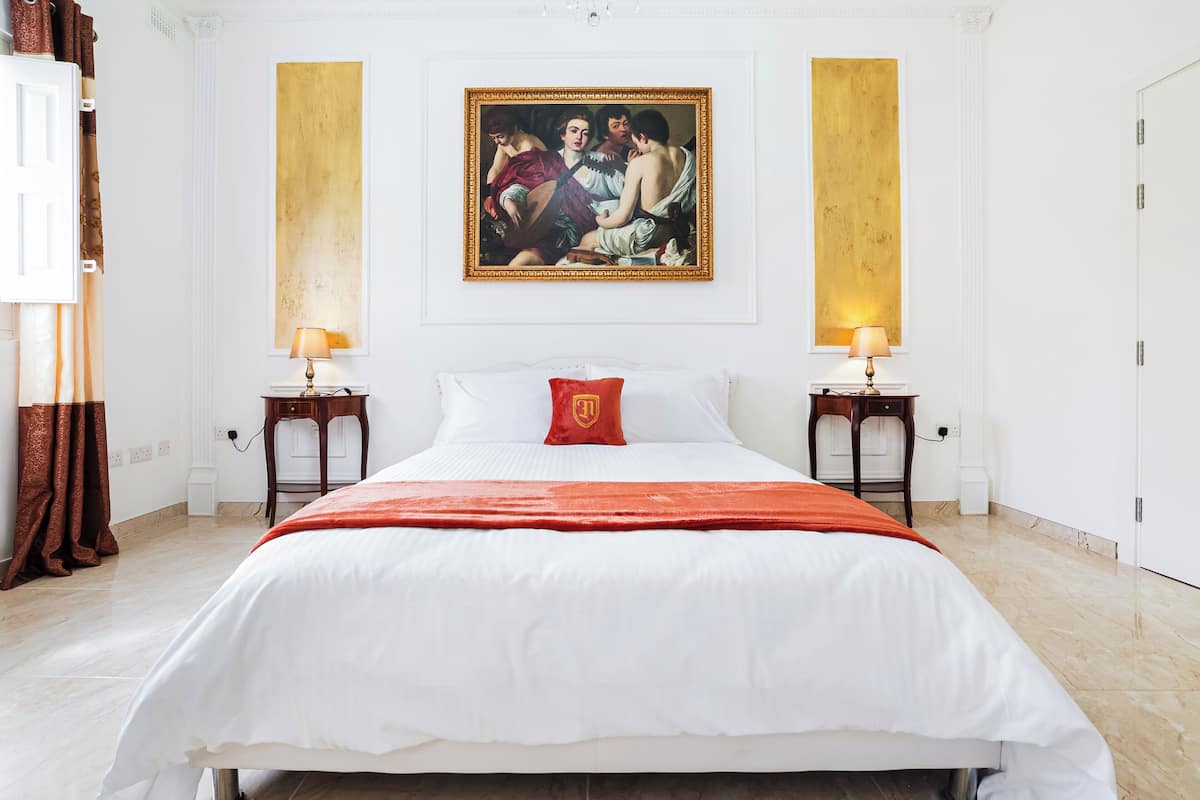 Une chambre d’hôtel avec un grand lit, deux tables de chevet, un oreiller rouge, un tableau au mur et une porte sur le côté droit.
