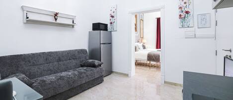 Een moderne woonkamer met een grijze bank, een kleine tafel, een televisie en een koelkast. Er is een gang die leidt naar een slaapkamer met een bed en een rood gordijn.
