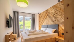 A bedroom with a wooden bed, a TV, a small wooden stool, and a window with curtains. - TOP 6 - Großzügiges Apartment mit Bergblick (Berwang)