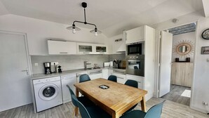 A modern kitchen with a dining area, including a wooden table and chairs, a washing machine, and a microwave. - Captain's Quarters - Maison en Bord de Plage (Saint-Côme-de-Fresné)