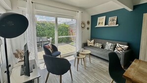 A modern living room with a grey sofa, a desk with a computer, and a dining table. There is a large window with a view of a garden and a blue wall with framed pictures. - Captain's Quarters - Maison en Bord de Plage (Saint-Côme-de-Fresné)