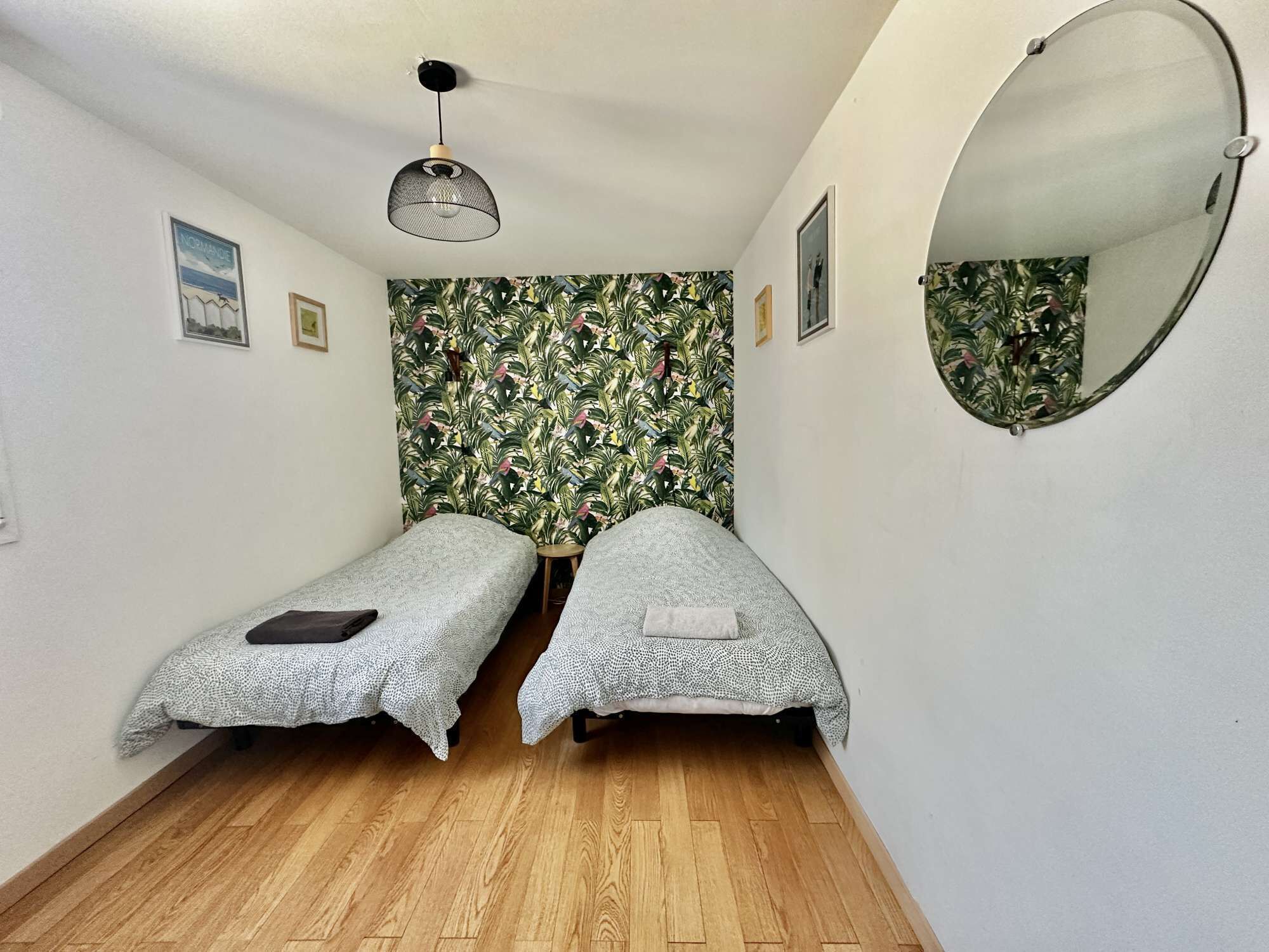 Um quarto pequeno com duas camas de solteiro, piso de madeira, espelho fixado na parede e luminária no teto.