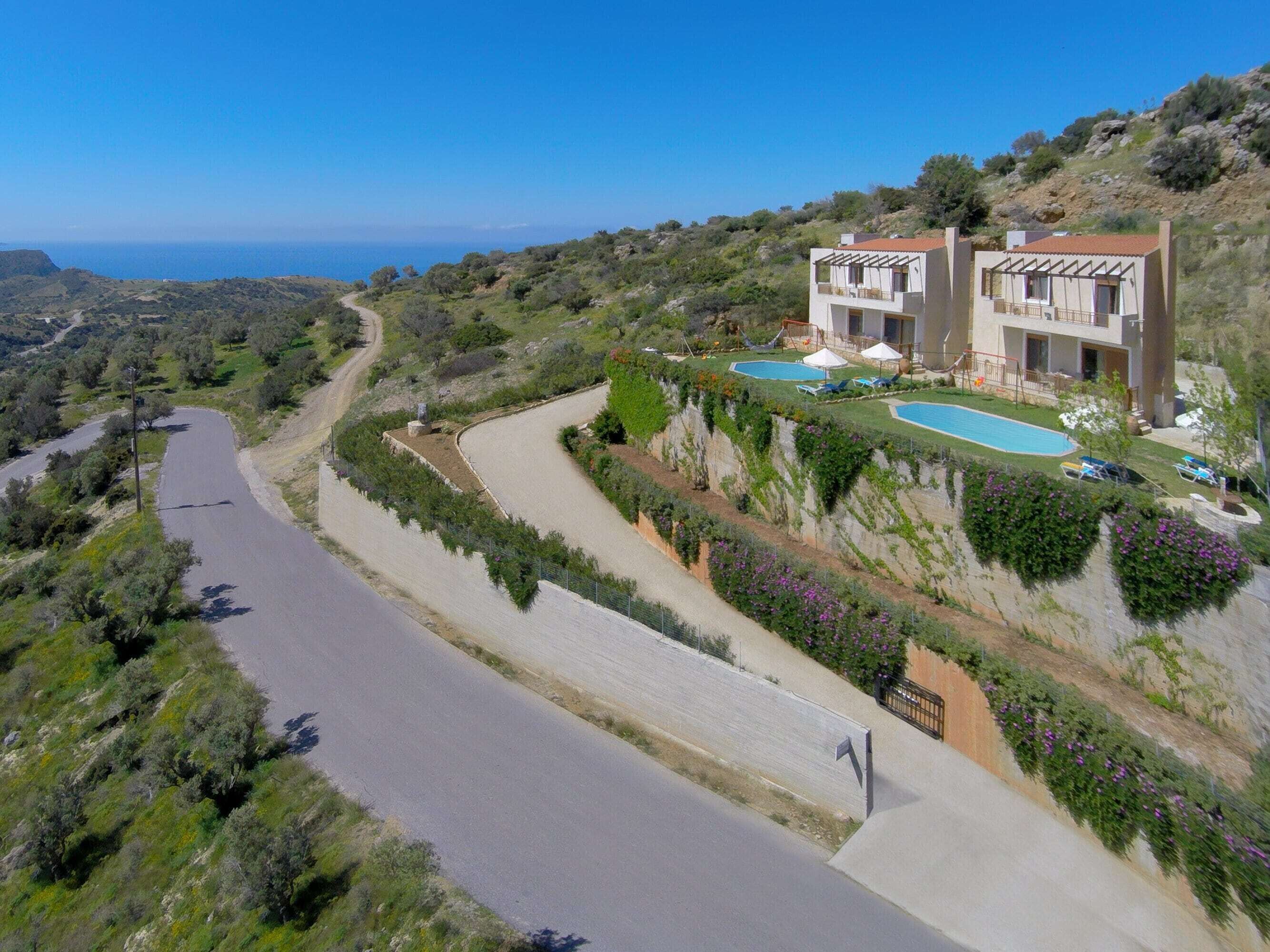 Um terreno em encosta com uma estrada sinuosa, duas casas modernas com varandas, uma piscina e vista para o mar.
