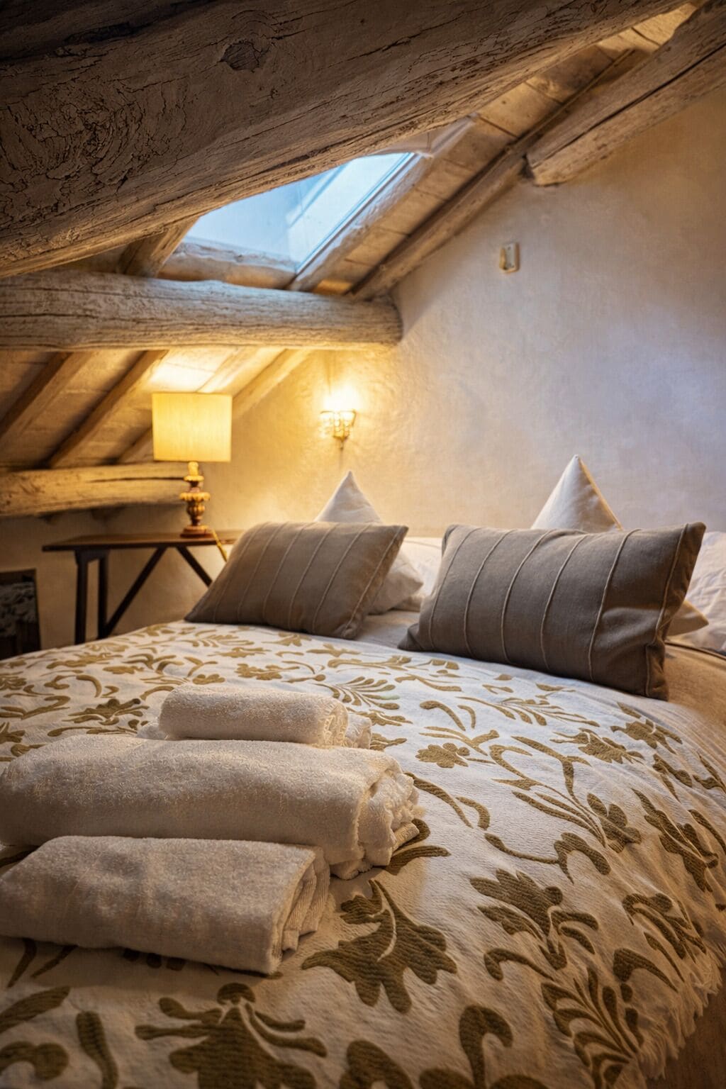 A cozy attic bedroom with a sloped ceiling, a skylight, a bed with a floral bedspread, and a bedside lamp.