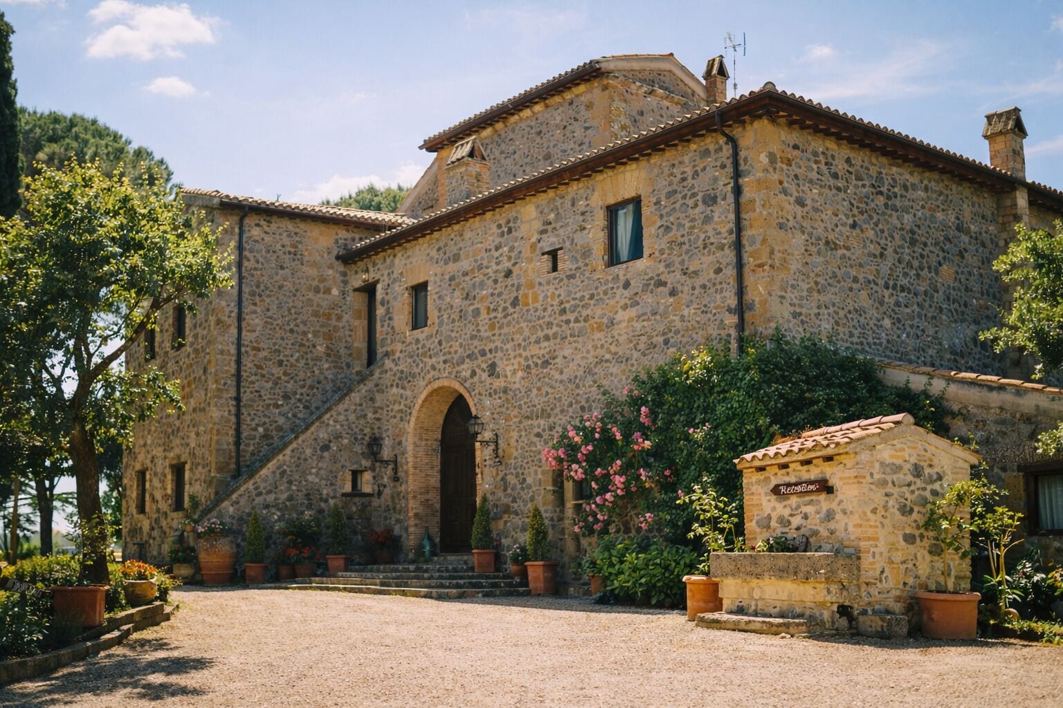 A stone building with a central arched entrance, a small annex with a sign, and a gravel driveway.