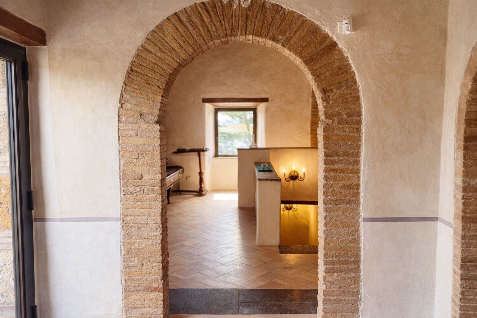 Uma porta em arco dá acesso a um quarto com chão em padrão herringbone, uma janela e uma luminária fixada na parede.