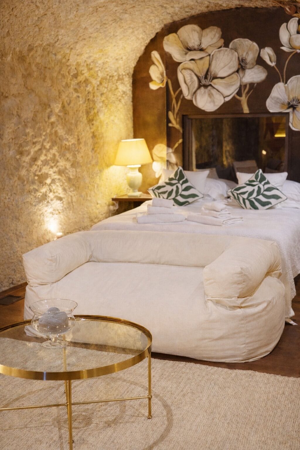 Un dormitorio con una cama grande, un sofá, una mesa de vidrio con una vela y un mural floral en la pared.