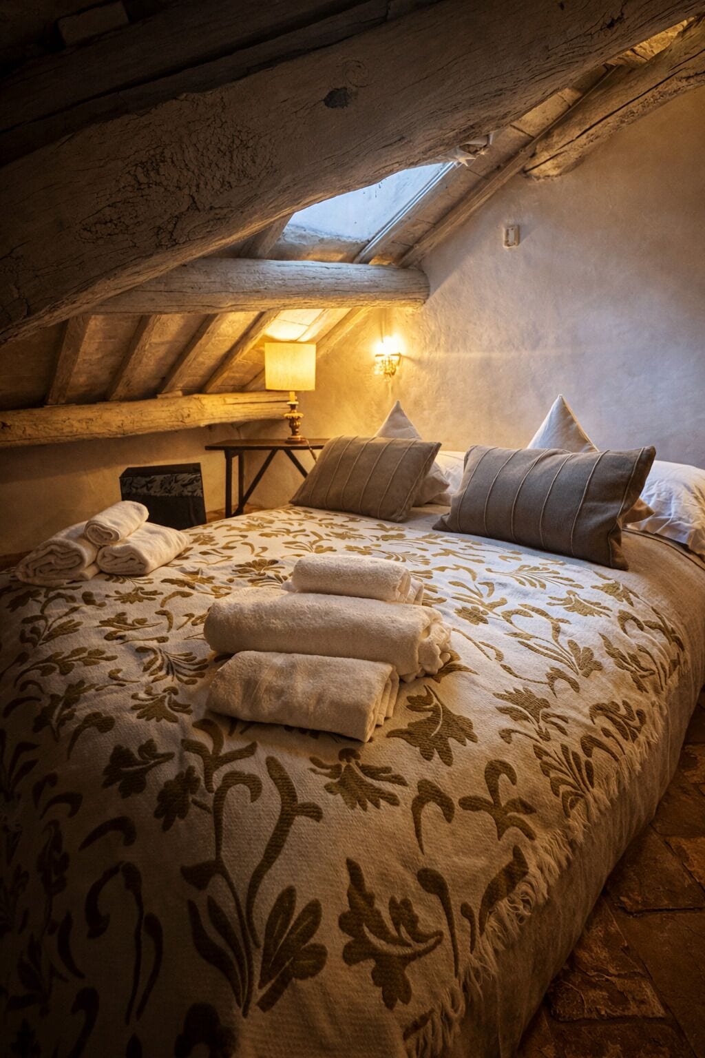A cozy attic bedroom with a sloped ceiling, a skylight, a bed with floral bedding, and a bedside lamp.