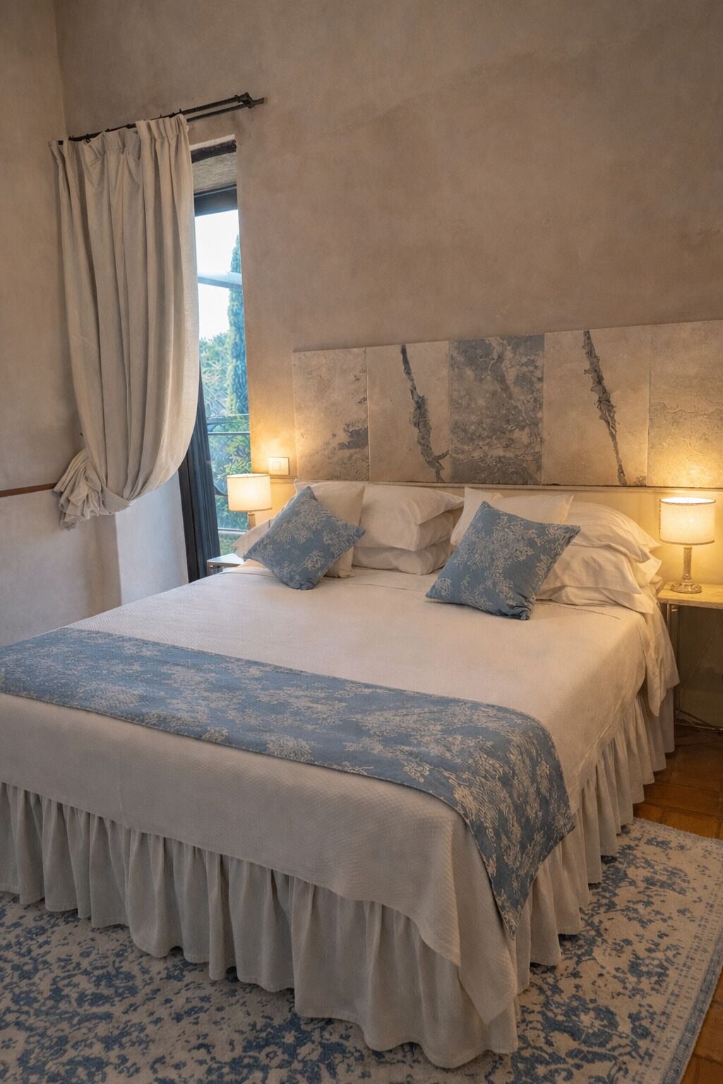 Une chambre à coucher avec un grand lit, deux lampes de chevet, une tête de lit au motif texturé et une fenêtre avec des rideaux.