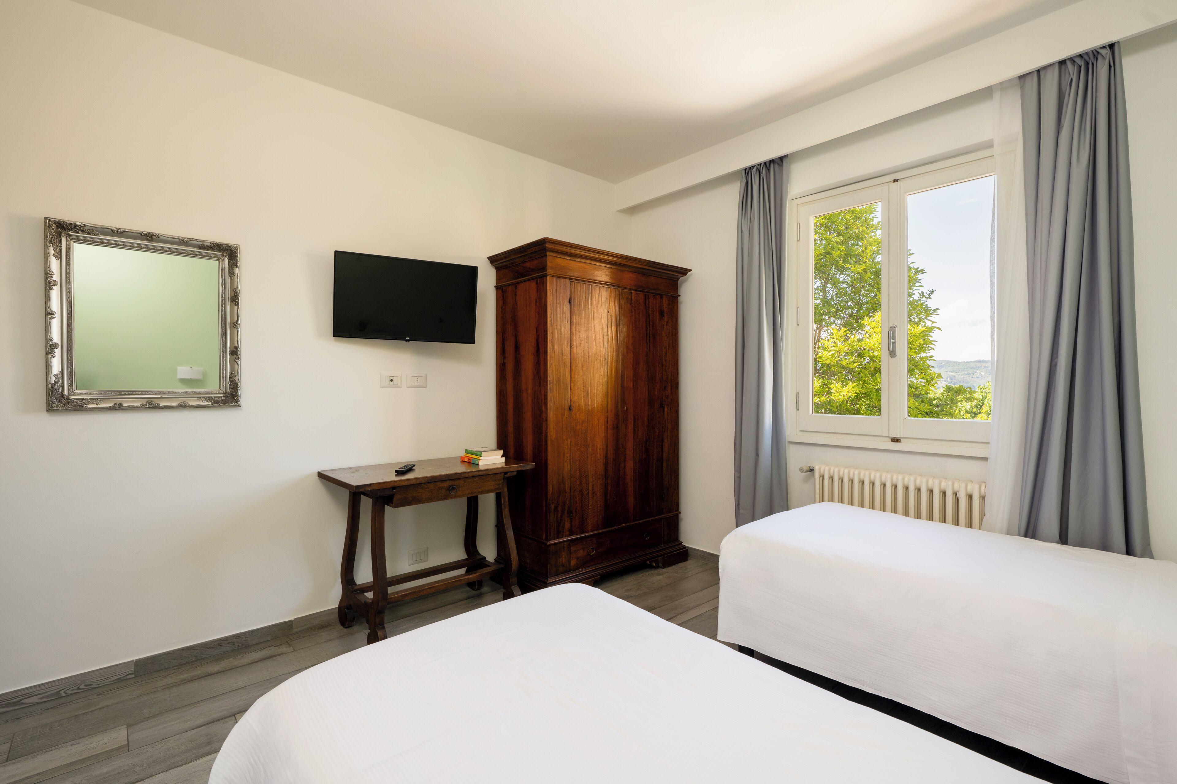 A hotel room with two beds, a wooden wardrobe, a TV mounted on the wall, and a window with curtains.