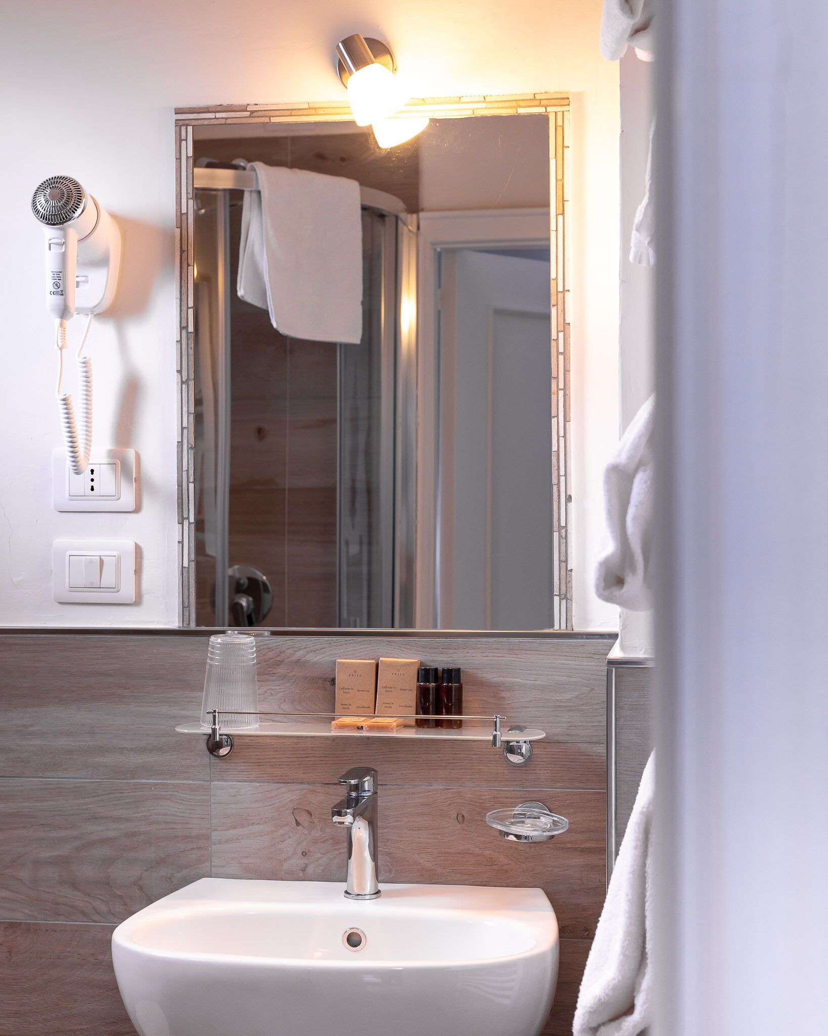 Un baño con lavamanos blanco, espejo, secador de pelo y artículos de aseo en un estante.