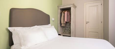 A bed with white bedding, a wooden nightstand, a wardrobe with hanging clothes, and a door.