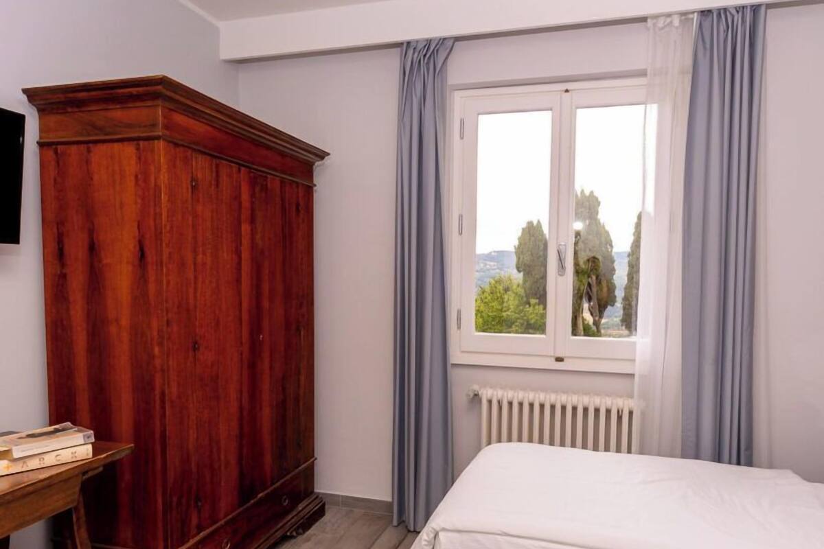 A bedroom with a wooden wardrobe, a bed, a window with curtains, and a radiator.
