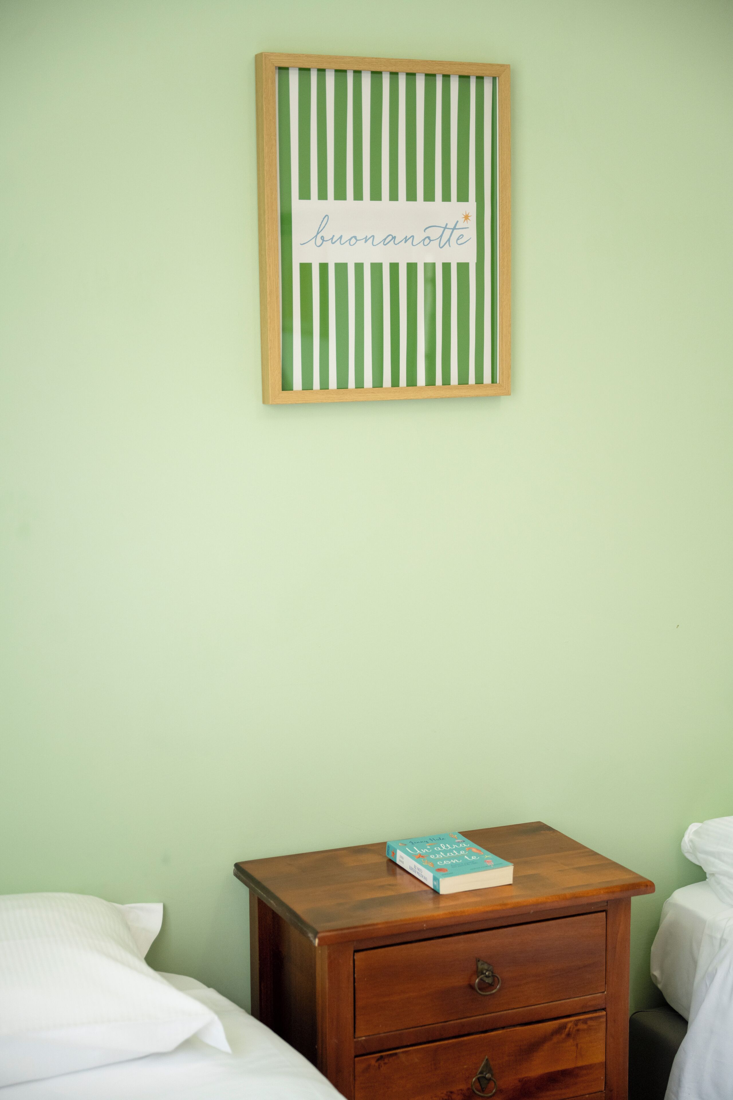 Un dormitorio con una cama, una mesita de noche con un libro y un cartel enmarcado en la pared.