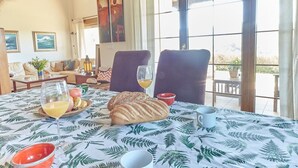 A dining table with a fern leaf tablecloth, two chairs, a loaf of bread, a glass of orange juice, and a bowl of fruit. - Home in Comillas · 3 bedrooms · 4 beds · 3 baths (Ruiloba)