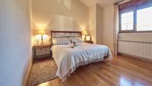 A bedroom with a wooden floor, a bed with a white bedspread, two bedside tables with lamps, a window with curtains, and a radiator. - Home in Comillas · 3 bedrooms · 4 beds · 3 baths (Ruiloba)