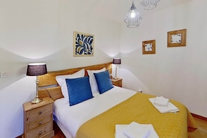 Ein Bett mit Holz-Kopfstück, zwei Nachttische mit Lampen, eine gelbe Tagesdecke und Kunstwerke an den Wänden.
