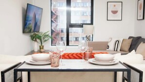 A dining table set with plates, glasses, and a bowl in a modern room with a large window, a sofa, and framed artwork on the walls. - City Centre Apartment, Quiet and Cozy, Sleeps 4. (Liverpool)