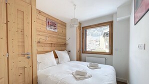 A bedroom with a wooden headboard, a large window, and a bed with white bedding. - Ambre blanche - Les Gets - Centre village (Les Gets)