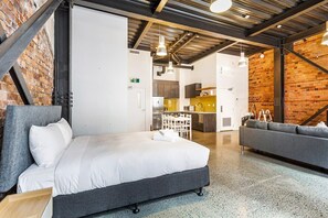 Moderni hotellihuone, jossa on suuri sänky, sohva, pieni keittiö ja paljaat tiiliseinät.