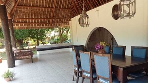 Un área de comedor al aire libre con una mesa y sillas de madera, techo de paja y faroles colgantes.