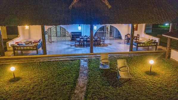 A thatched-roof outdoor dining area with tables and chairs, surrounded by white walls and a covered seating area.
