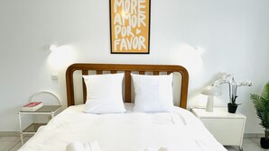 Una cama bien hecha con sábanas y almohadas blancas, una mesita de noche con un libro y una lámpara, y un cartel enmarcado sobre la cama.