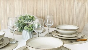Una mesa de comedor dispuesta con platos, cuencos y copas de vino, junto a una pequeña planta en maceta.