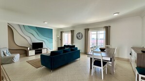 A modern living room with a blue sofa, a TV on a stand, a dining table with chairs, and a large window with curtains.