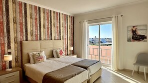 Et hotelværelse med en stor seng, udsigt over byen fra en balkon og et maleri på væggen.