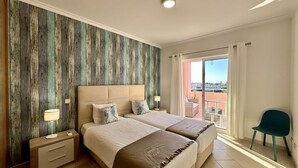 A double bed with a wooden headboard, two bedside tables with lamps, a view of the city through a balcony, and a green chair.